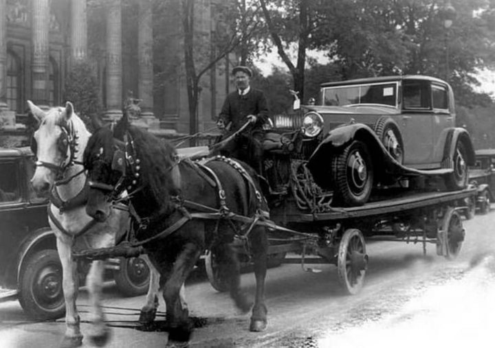 A 'period' classics pictures thread (Mk III) - Page 308 - Classic Cars and Yesterday's Heroes - PistonHeads UK - The image appears to be a black and white photograph. It depicts a scene from the early 20th century featuring an old-fashioned car being transported on a trailer by two horses. The horses are harnessed to a wagon, which is pulling the trailer with the car on it. In the background, there's a man riding on the trailer, likely overseeing the transportation process. The setting suggests a city street, given the presence of cars and buildings in the background.