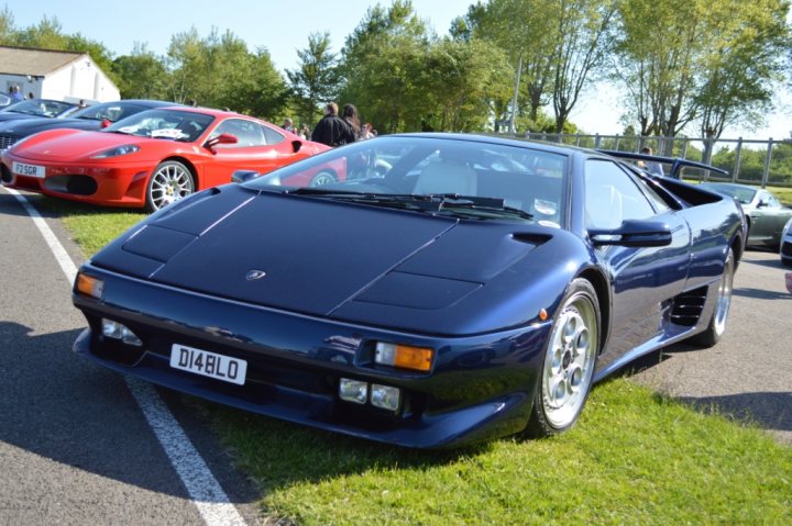 Real Good Number Plates : Vol 4 - Page 58 - General Gassing - PistonHeads - The image showcases a row of luxury cars parked on a grassy field, with a prominently featured blue Lamborghini Diablo sports car in the foreground. The car is sleek and streamlined, with a shiny blue exterior. A red Ferrari is visible in the background, adding to the array of high-end vehicles. There are also people scattered around the cars, possibly owners or enthusiasts. The backdrop consists of a simple white building and a fence, providing a contrast to the luxury cars. The grassy field suggests an outdoor car show or exhibition.