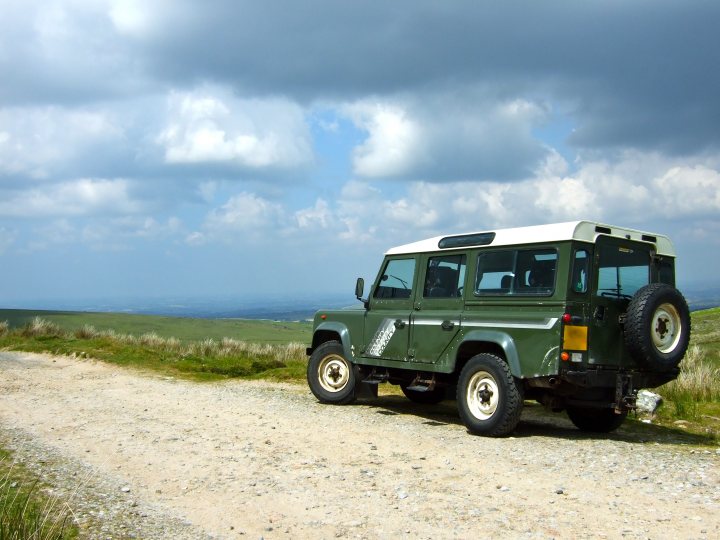 Land Rover Pistonheads - In the image, a green and white Land Rover is parked on a gravel road. The vehicle is angled slightly towards the camera, showing its robust design and four-wheel drive capability. The presence of mountains in the background adds a sense of ruggedness to the scene. Above, the sky is filled with clouds, suggesting the possibility of rain or impending storm. The Land Rover's position on the gravel road suggests it might be used for off-road driving or exploring the wilderness areas.