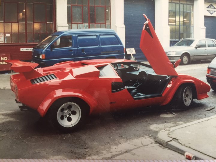 My old Lambo photos from the 90s - Page 21 - Lamborghini Classics - PistonHeads - The image presents a striking red convertible parked on a city street. The car's roof is open, with the interior unveiled as inviting as a summer day. Around the car, you can see typical elements of an urban setting: a blue van parked to the left, a gray saloon car to the right, and a brick building featuring large windows in the background. A white sign is also visible to the left of the frame. The colors, objects, and layout in the image suggest a bustling city atmosphere, perhaps during a sunny day.