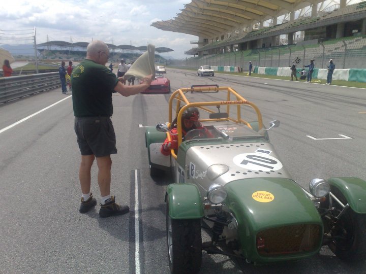 Sepang Afos Pistonheads - The image captures a scene at a racing track. A man is standing by a green car, which appears to be an older model with a distinctive number 10 on its side. The car is parked at a garage stall, suggesting a time of pause or preparation in the race activity. In the background, various people are scattered across the scene, possibly staff or other participants at the event. The presence of several cars in the background implies that this is a busy and active environment. The structures, which include multiple benches, indicate that the track is equipped with facilities for spectators or resting participants.