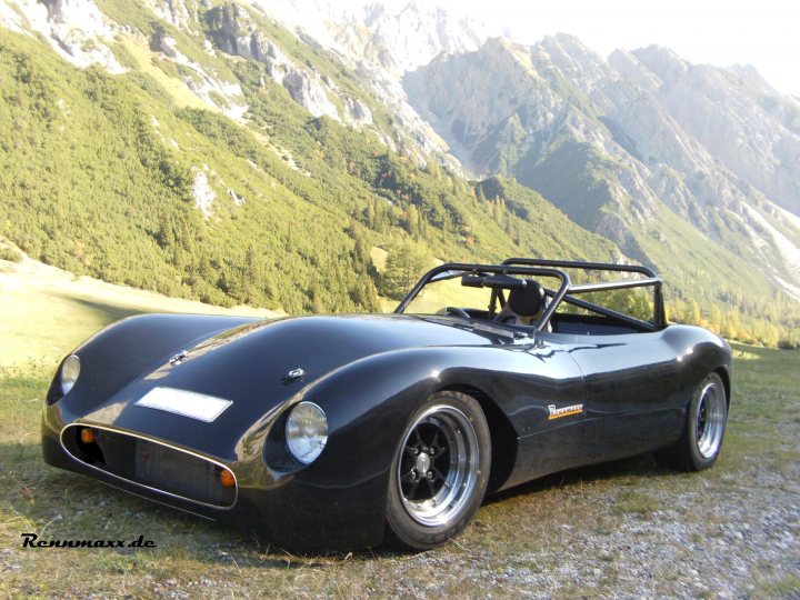 The Best looking Kit Cars - Page 9 - Kit Cars - PistonHeads - The image features a sleek, black sports car resting on a grassy hillside. The car is positioned prominently in the foreground, its curved shape accentuated by the landscape. Behind the car, an expansive valley unfolds, with a range of mountains towering in the distance under a clear blue sky. The lighting suggests it's early morning or late afternoon, casting soft shadows and lending a serene atmosphere to the scene. The car's reflective surface seems to capture the light, enhancing its streamlined design.