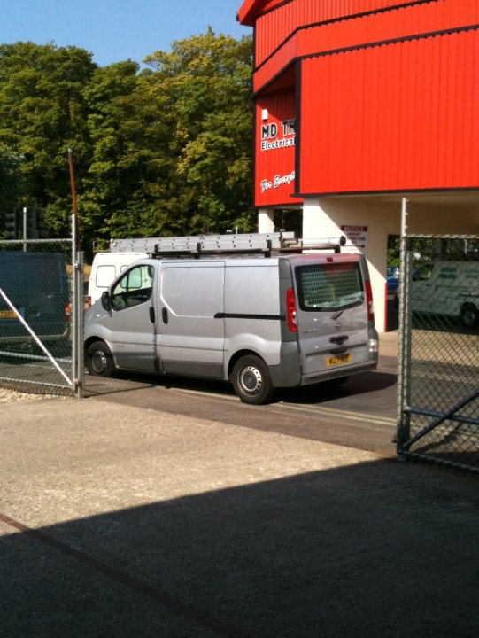 Bad Pistonheads Thread Parking - The image shows a gray van parked next to a utilities worker, likely on a break. In the background, there's a red wooden building with metal elements and a red door, which could be a company structure. A chain-link fence is visible on the left, suggesting a secure industrial or commercial area. The sky is clear and blue, indicating a sunny day.