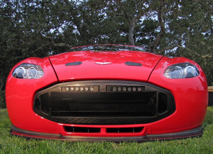 Pics from Aston event, Monterey - Page 1 - Aston Martin - PistonHeads - The image features a red convertible car. The car's convertible top appears to be closed, and the vehicle is parked on grass, angled slightly to reveal the front grill. The grill design is distinctive with a pattern that suggests the car might be an Audi. The hood of the car has an emblem on it, commonly known as an "Audi badge." The car is all-red with no visible customizations or modifications, and it stands out against the less vibrant background of a grassy lawn with trees in the distance.