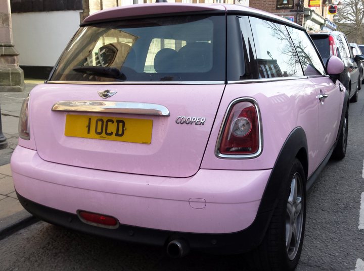 Real Good Number Plates : Vol 4 - Page 311 - General Gassing - PistonHeads - The image shows a pink Mini Cooper car parked on a city street. The car has a British-style license plate mounted on the back. There is a clear glass window at the back, providing visibility into the car. The opposing side of the street includes parked vehicles, a building, and a lamppost, suggesting an urban setting. The image is slightly blurry, hinting at a moving vehicle, possibly the Mini Cooper itself.