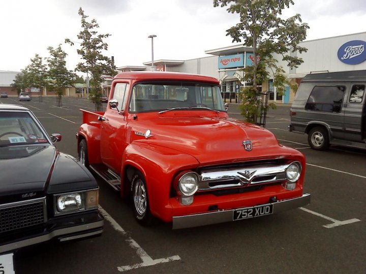 Cruisemeet Pistonheads Edinburgh - The image captures a parking lot scene that includes several vehicles. In the forefront, there is a bright red vintage truck parked next to a black car. The background shows other parked vehicles, including another car and a minivan. A store called "Pizza Hut" is visible in the far background, suggesting that this location may be frequented by customers for dining or takeout. The setting suggests a casual, everyday environment, potentially taken on a cloudy day.