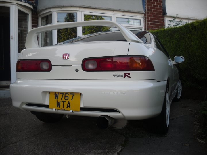 Pistonheads - This image captures a white Honda RX-R sports car parked on a driveway, featuring a striking red taillight design above a yellow and black license plate. The car is parked in front of a house, with a portion of the hedge visible, enhancing the residential setting. The vehicle is angled towards the left side of the frame. The overall scene suggests a quiet suburban neighborhood.