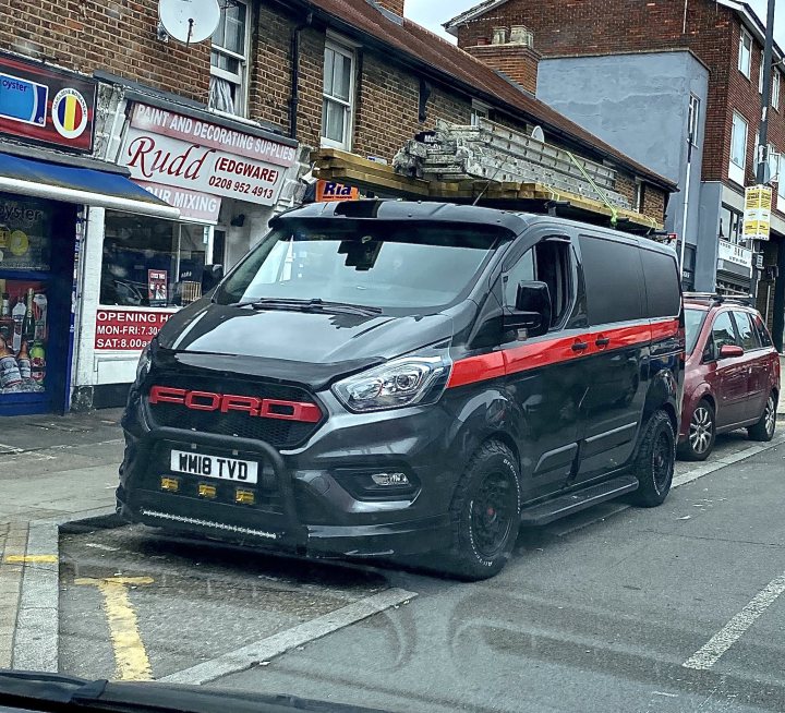 Pictures of decently Modified cars [Vol. 2] - Page 402 - General Gassing - PistonHeads - The image features a distinctive black and red Ford van parked on the side of a street. The vehicle has a large, elevated platform at the back, which appears to be used for transporting items. It's equipped with a black roller door and black wheels. There is text on the van that reads "Ford" and "Ranger," indicating its make and model. In the background, there are shops and buildings, suggesting an urban setting. The vehicle's position on the street indicates it is likely parked for loading or unloading goods, given the platform at the back.