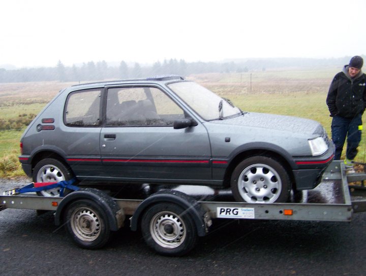 205 gti gti-6 - Page 1 - French Bred - PistonHeads - The image shows a grey hatchback car being transported on a flatbed trailer in what appears to be rainy weather. The car is positioned on the trailer, supported by four wheels on a metal platform, facing towards the right side of the image. The car's red stripe along the side and the black roof contrast with its grey body. A person, partially visible on the right side of the image, is standing next to the car, observing the operation. The cloudy sky and overcast conditions suggest the weather is less than ideal. The overall scene suggests a situation where the car might be experiencing mechanical issues and is being towed.