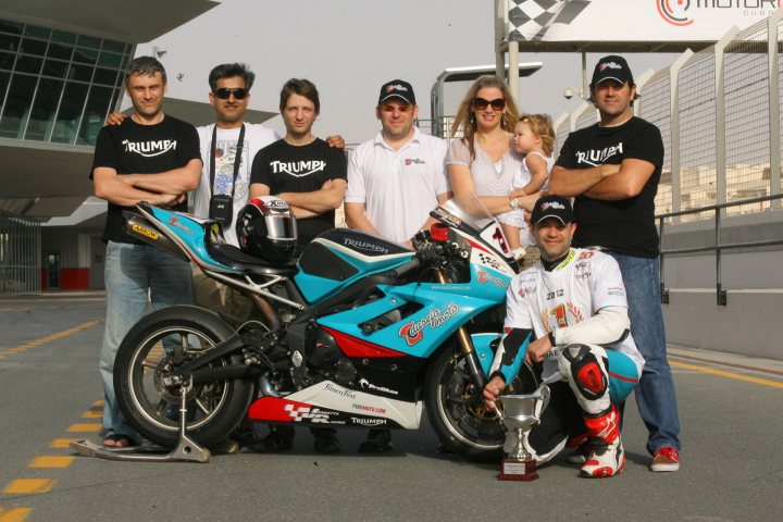 Pistonheads - This is a color photograph featuring a group of eight individuals standing behind a blue and red motorcycle. They are posing with smiles, some of them wearing matching black "TRIUMP" shirts, which suggests they may be part of a racing team or event. The motorcycle is adorned with racing livery, including the same "TRIUMP" branding on the front and the number 22, indicating its involvement in a sport or competition. The setting appears to be a racetrack or similar motorsport venue, as indicated by the checkered flags in the background. This scene conveys a sense of camaraderie and celebration, likely in connection with a victory or significant achievement in the world of motorcycle racing.