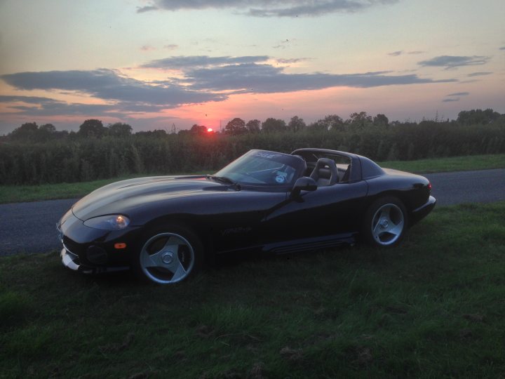 i phone pic - Page 1 - Vipers - PistonHeads - The image showcases a distinctive black sports car parked on the grass alongside a road. The car, with its convertible top and open side windows, exudes a sense of speed and freedom, accentuated by the setting sun in the background. The field and trees in the distance add a serene backdrop to the sports car, creating a striking contrast between the dynamic nature of the vehicle and the tranquil rural landscape. This scene is a captivating depiction of the blending of human-made technology with the beauty of nature.