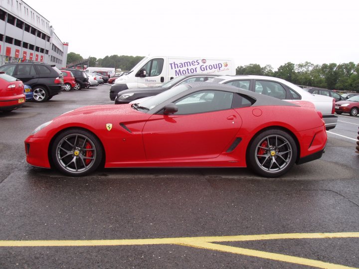 Pistonheads - The image shows a close-up of a striking red Ferrari sports car parked in an outdoor lot. The car is positioned diagonally, focusing on the front and side. It appears to be a convertible model with a sleek design and distinctive features such as the large air intake at the back and the characteristic yellow Ferrari emblem. The background is filled with other cars and trucks, suggesting that this location might be a public parking area or a showroom. The lighting and shadows on the ground hint at it being either early morning or late afternoon with overcast skies.