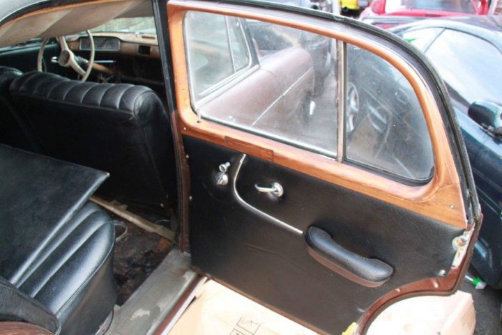 1950's Merc 220s Saved from the grave - rat rod restoration  - Page 1 - Mercedes - PistonHeads - This image provides a unique perspective from inside an old car's driver's side door. The open door offers a glimpse of the vehicle's interior, which includes a classic leather-upholstered seat matching the door's material, a wooden arm rest, and a steering wheel that is not visible. Beyond the open door, visible traffic lights suggest that the car is parked in a city or urban environment.