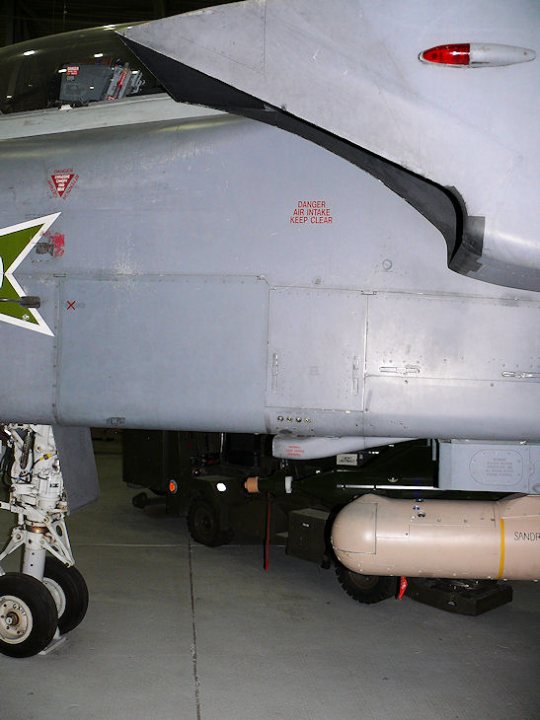 An airplane is parked on the tarmac at an airport - Pistonheads - The image displays the underside of what appears to be a jet aircraft. The jet is predominantly grey with some visible components, such as the engine, situated below the body. The visible area includes a text inscription which is partially obscured and illegible. There are also various markings and identifiers on the body of the jet. Additionally, a red reflector or light is visible, suggesting safety or navigation features. The background is nondescript and appears to be a maintenance or storage facility, indicated by the presence of additional metres of the aircraft.