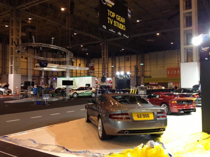 Top Gear Live (NEC this weekend) - Page 1 - Aston Martin - PistonHeads - The image captures an indoor car show or exhibition. There are various cars displayed in a large warehouse or hangar. A grey Aston Martin is prominently featured, with a yellow license plate. The background features a television and a sign that reads "TOP GEAR TV STUDIO." There are other cars and screens in the background, suggesting a bustling event or exhibition. The overall atmosphere is one of a professional car show or event.