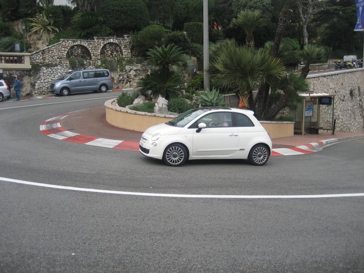 Pistonheads Week Carlo Monte - The image captures a moment on a winding street, where a small white Fiat car is making a right turn. The Fiat is the central figure in the scene, situated on the left side of the image. The road itself is a testament to the area's terrain, winding its way through the landscape.

In the background, the street gives way to nature, as it is bordered by lush trees and bushes on the right side of the image. These green companions, standing tall, add a touch of tranquility to the otherwise urban scene.

Just off the main street, a sidewalk runs parallel to it, providing a safer path for pedestrians. It contrasts with the road, which is dedicated to vehicles. A few people can be spotted on the sidewalk, engrossed in their own worlds and adding life to the otherwise still image.

Lastly, the presence of a building on the right side of the image signifies that this street is part of a residential or commercial area. It's a snapshot of an everyday scene, filled with life and activity.