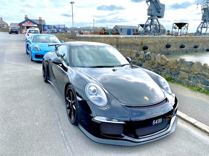 Show off your GT, past and present... - Page 53 - 911/Carrera GT - PistonHeads - The image shows a black sports car parked on the side of a street. It's positioned in front of other cars, suggesting it might be at an event or a gathering. The setting appears to be a coastal town with a marina visible in the background, under a cloudy sky. There are no people immediately around the car, and the focus is on the vehicle itself, which has a shiny finish reflecting some of the light.