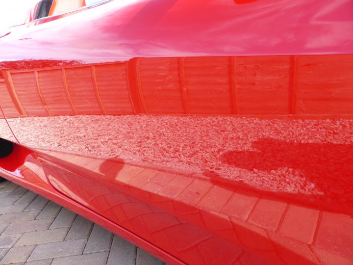 Car valeting full detail in South Devon - Any recommendation - Page 1 - South West - PistonHeads - The image displays a close-up of the side and rear of a bright red car. The car appears to be parked, and its glossy red paint reflects the city street where it's located. There is a clear, refined texture visible on the car's surface, suggesting a high-quality finish. The image effectively captures the car's presence, accentuating its vivid color and shiny finish. The photograph has a focus on the car's exterior, highlighting its sleek design and polished surface.