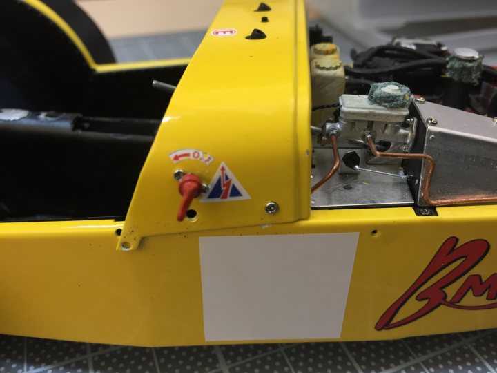 Tamiya Caterham 1/12 - modified - Page 1 - Scale Models - PistonHeads - The image shows a close-up view of the back end of a kart, focusing on its structure and design. The kart is predominantly black, but the part visible in the photo is painted yellow. The name "B&M" is visible on the back bumper of the kart, indicating a possible brand or team affiliation. There are several bolts visible, suggesting the internal components are attached to the exterior shell. The kart appears to be in a work or storage environment, as indicated by the protective surface beneath it and the clutter in the background.