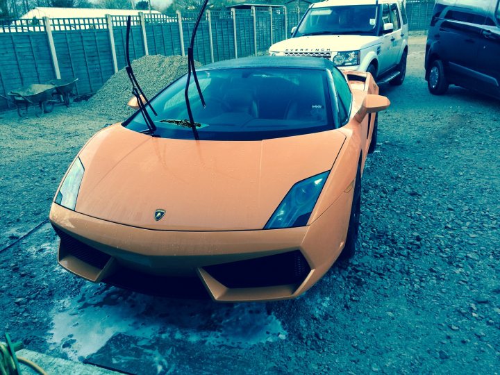 What is my colour code ? - Page 1 - Gallardo/Huracan - PistonHeads - The image features a vibrant orange Lamborghini parked on a gravel surface. The car's unique design and shiny finish are clearly visible. In the background, there is another car and some vehicles, suggesting a parking area or a gathering of some sort. The sky above is overcast, casting a soft light that seems to reflect off the car's surfaces. There are some objects on the ground near the car, possibly pieces of debris or personal belongings.