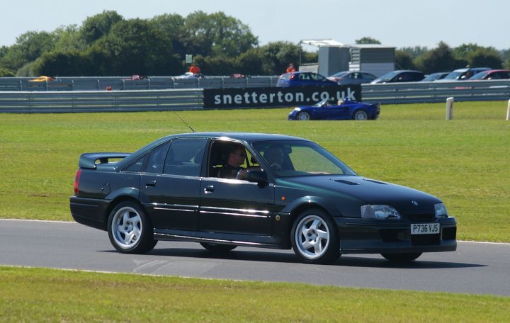 Snetterton Lotus Festival 25/26 June - Page 1 - General Lotus Stuff - PistonHeads - The image depicts a black two-door car racing on a track with grassy areas and trees along the circuit. Another blue car is present in the background, suggesting a race or test event. There is a visible logo displaying "Snetterton.uk," indicating the location or the track, and a car by the same name, perhaps a sponsor or the racing team. The scene conveys a sense of speed and competition characteristic of a motorsports event.