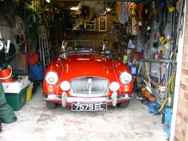 Winter Shed Pistonheads - The image showcases a garage scene with a vintage red sports car as the central focus. The car is positioned at an angle, revealing its chrome grille and shiny exterior. The garage itself is filled with an array of household items, including bicycles, various tools, and an empty wall covered with an assortment of items. The floor is covered in dirt and tools, suggesting recent use or perhaps ongoing maintenance. Across from the car, leading into the room, is a white door. The overall setting suggests a well-used garage space filled with both automotive and day-to-day items.