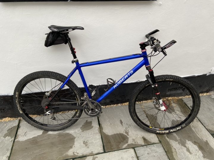 The "Show off your bike" thread! (Vol 2) - Page 7 - Pedal Powered - PistonHeads - This is a photograph of a blue mountain bike parked against a white wall with a black border at the bottom. The bike has a number of features including a front suspension fork, disc brakes, and is equipped with knobby tires suitable for off-road use. It also has a water bottle holder and a bell, suggesting it might be used for cycling in urban or rural environments. The bike's frame appears to have some visible dirt, indicating recent use. There is no text present on the image.