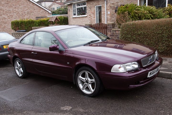 Volvo c70 t5 turbo help - Page 1 - Volvo - PistonHeads - This image features a dark purple car parked on a street. The vehicle is a compact, four-door sedan with polished alloy wheels, and it's parked in front of a brick house with a well-maintained garden. To the left of the image, there's a glimpse of another vehicle, partially obscured. The setting appears to be a residential area, as suggested by the house in the background. No people are visible in the photo.