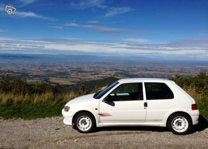 Peugeot 106 Rallye - I am going to look at... - Page 1 - French Bred - PistonHeads - The image presents a picturesque scene from atop a hill. At the center of the frame is a small white car, parked on a gravel road. The car, surrounded by the serene landscape, adds a sense of scale to the vast, open area. The road leads to a cliff edge, from where the view falls away into an expansive valley below. Further in the distance, a small white patch on the sky stands out against an otherwise clear sky, adding a touch of mystery to the scene.