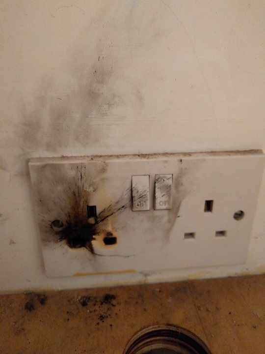 Burnt out plug / socket advice - Page 1 - Homes, Gardens and DIY - PistonHeads - This image shows a close-up of an electric outlet with evidence of fire damage. The outlet is white and appears to have been previously used, with sparks indicating a short circuit or an electrical incident. There is a melted area around the outlet, which suggests that the fire caused some destruction. In the surrounding space, there is a visible presence of soot and burned debris. The image conveys a sense of urgency and the need for repair or replacement.