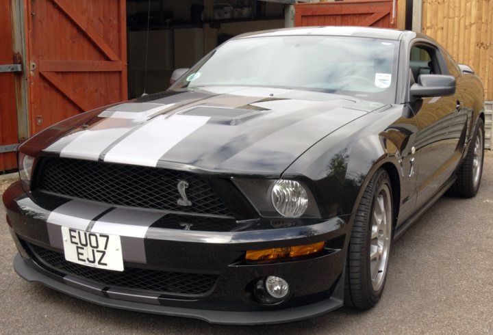 My new Mustang acquisition... - Page 1 - Mustangs - PistonHeads - The image depicts a black Mustang car with a glossy finish and silver detailing along the hood and the side. The car has a European license plate that reads "EU07 EJZ," indicating it might be registered in a European country or it could be owned by someone living in Europe. Behind the Mustang is a wooden structure, possibly a barn or stable. In the background, there is another vehicle that is partially visible. The overall scene suggests that the Mustang is parked or moving slowly on a paved surface.