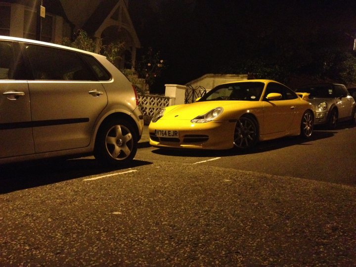 A car is parked on the side of the road - Pistonheads - The image captures a peaceful scene of two cars parked on what appears to be a residential street. The first car is a silver hatchback, parked close to the curb. The second car is a vibrant yellow sports car, parked on the other side of the street. Both cars are parked in front of a white house, which features a visible garage door. The setting appears to be late in the evening, as indicated by the diminished visibility and the dark sky. The overall image conveys a sense of tranquility and order.