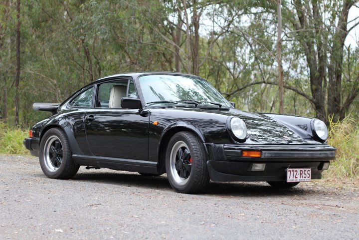 Pistonheads - The image features a black Porsche 911, specifically a GT2 RS model, parked on a gravel surface surrounded by a lush green forest. The car is positioned at an angle, with the front left half visible, emphasizing its distinct nose and curves. The license plate reads "772 RSS", indicating that it is a show car. The setting appears serene and calm, with the gravel and greenery contrasting against the sleek black exterior of the car. The image captures the elegance and power of the Porsche in a tranquil natural environment, a scene that mixes the beauty of nature with the perfection of automotive design.