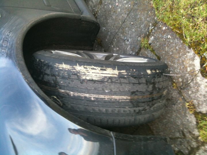 Don Palmer car control course - Page 1 - Advanced Driving - PistonHeads - The image shows a flat tire on a vehicle, with the wheel retracted into the hubcap. There is visible tire damage, with extensive and uneven wear on the tire. The tire tread pattern appears mostly worn away, indicating significant use. The tire is leaning to one side, which is typical after a flat tire incident. The surrounding ground is covered with grass and gravel, suggesting a driveway or a similar outdoor setting.