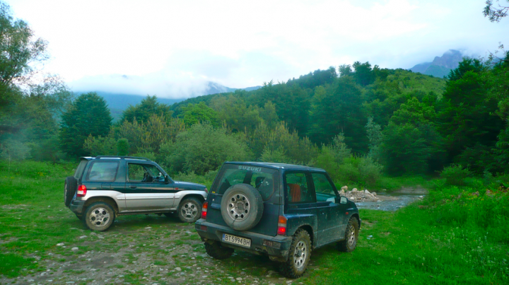 Little overnight trip to the Balkan range - Page 1 - Off Road - PistonHeads - The image captures a tranquil woodland scene, featuring two vehicles parked within a grassy clearing. A small jeep, characterized by its rugged black color and spare tire mounted on the back, sits closer to the viewer. The larger vehicle, painted in shades of green and white with a similar spare tire design, is parked slightly further away. Both vehicles are facing in the same direction, as if ready to embark on a journey through the verdant landscape. The backdrop teems with lush green trees and a distant mountain range, lending a sense of adventure and natural beauty to the scene.