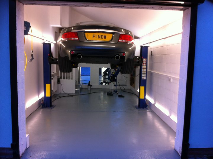 Garage Queens - Page 10 - Aston Martin - PistonHeads - The image captures a silver car inside a car lift. The lift, supported by yellow and blue poles, is lifting the back end of the vehicle. The car's license plate reads "FINDING," and it's positioned above a small car lift. The setting appears to be a garage or maintenance workshop, and there is a single person present, possibly a technician or mechanic. The lighting suggests it might be nighttime, as the area has a dim ambiance. The ceiling shows signs of heavy machinery use, indicating that this is a space dedicated to automotive tasks.