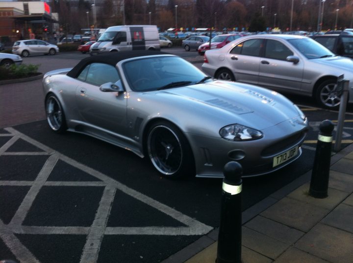 Pistonheads - The image depicts a modern, two-door sports car parked at an angle on a parking lot. The car is silver in color with a sleek design and is positioned in front of a row of parking posts. In the immediate vicinity of the car, there's another vehicle that appears to be a silver sedan, partially visible. The background reveals a busy parking lot with several cars parked in rows. The setting suggests it might be a public parking area, possibly near a shopping center or commercial area. There are no visible texts or distinctive logos that provide additional information about the location or the car itself. The picture is captured during the day in natural lighting, which casts soft shadows and enhances the metallic sheen of the vehicle.