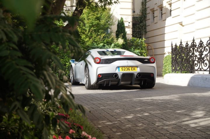 Unique 458 speciale aperta - Page 1 - Ferrari V8 - PistonHeads - The image features a white Ferrari parked on a sidewalk, its license visible. The car is set against a backdrop of a white building and lush green trees, casting a classic shadow on the cobblestone street. The Ferrari brand logo is prominent on the back of the car, indicating its luxury and exclusivity. A small red lamp is also visible, adding a pop of color to the scene. The setting appears to be a quiet, urban area, perhaps a residential neighborhood, given the presence of the Ferrari on the side of the street.