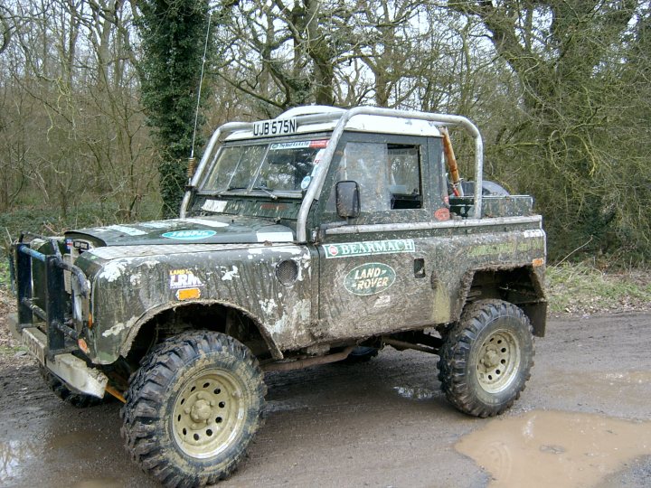 Went to the PN meet at the Ace last night... - Page 1 - Off Road - PistonHeads - The image shows a rugged off-road vehicle, which appears to be a well-used pickup truck. The truck has a green base color with mud splatters throughout, suggesting it was driven on a muddy trail or in wet conditions. The vehicle is equipped with large off-road tires suitable for rough terrain. Above the license plate, a roll bar can be seen for safety, and the door has a window that is not completely covered by glass. The truck also has a vintage radio on the dashboard. The surroundings include a forest with trees, giving the impression that the photo was taken in a wooded or natural environment.