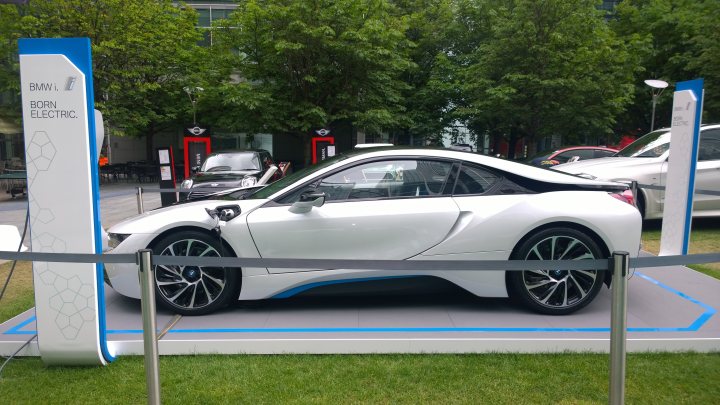 Tesla and i8 at Canary Wharf motorshow this week - Page 1 - EV and Alternative Fuels - PistonHeads - The image displays an electric car showcased in an outdoor setting. The car is white with blue highlights and is parked on a concrete foundation. A fence marks the boundary of the display area. In the background, there are other cars and a crowd of people, suggesting an event or festival. The greenery in the background adds a touch of nature to the urban exhibition.