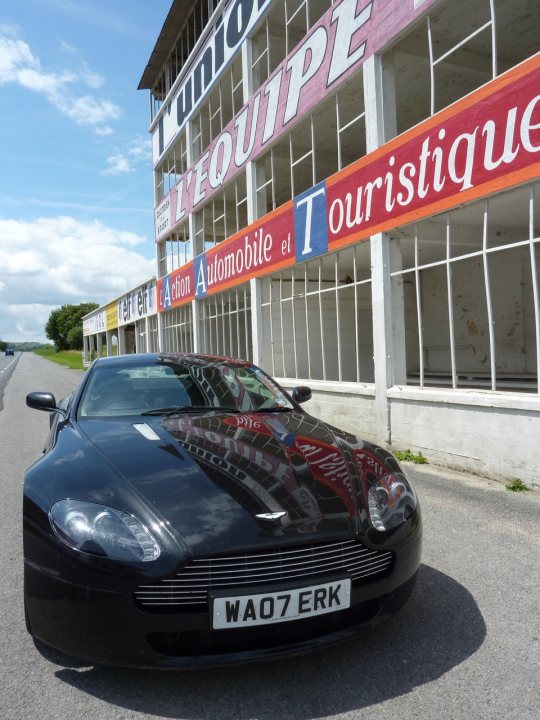 Trip of a Lifetime (picture heavy) - Page 1 - Aston Martin - PistonHeads - The image features a sleek black sports car parked in front of a building with a brightly colored sign that reads "Cercle Touristique Toursite." The signboard showcases text in both English and French, emphasizing tourist services. The perspective of the photo is from the side of the road, providing a clear view of the car and part of the building. The sky in the background is partly cloudy, suggesting outdoor weather conditions.