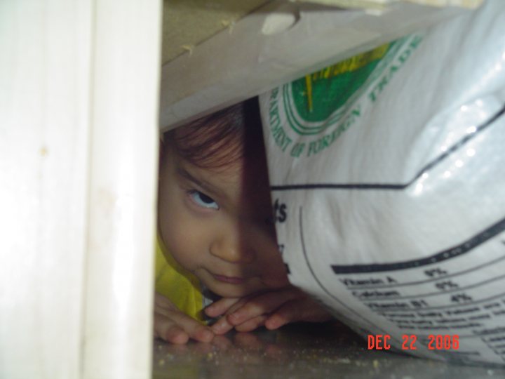 The image depicts a small child peeking out from under a bag, with the perspective of the camera positioned higher, perhaps to simulate the child's viewpoint. The bag is adorned with a logo that reads "Trade" which is likely the brand of the bag. There is a timestamp at the bottom right corner of the image, indicating it was taken on December 22, 2000. The tone of the image, with its low angle and shadowed corners, creates a sense of intrigue and playfulness.