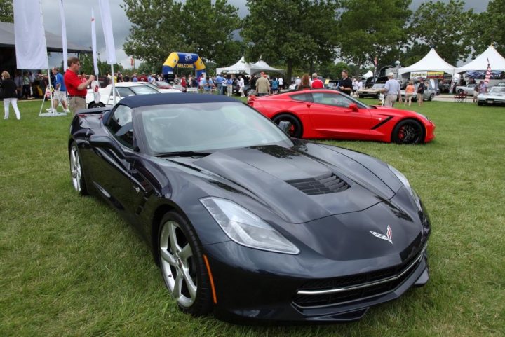 C7 spotted - Page 1 - Corvettes - PistonHeads - The image depicts a car show taking place in a park setting. There are two prominent sports cars on display: a black one and a vibrant red one. The cars are positioned on the grass, surrounded by spectators who are scattered around the exhibition area, and white poles that serve as banners. The background features a white tent and a cloudy sky, indicating an outdoor event under adventurous weather conditions. The vehicles appear to be connected to a monitoring system, one of which is showcasing an electronic dashboard and display, possibly indicating real-time performance data or inspection results. The image conveys a sense of luxury, showcasing high-end vehicles, and an atmosphere of anticipation and interest for the car enthusiasts present.