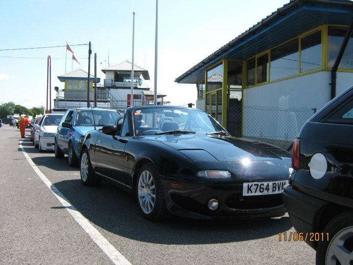 Pistonheads - The image portrays a bustling scene of a parking lot at a pier. The foreground is dominated by a row of sleek, black sports cars, each parked parallel to each other, their shiny exteriors gleaming under the sun. Behind the sports cars, there's a row of hatchbacks parked in the same orientation. The vehicles vary in color, including hues of white and blue. This arrangement suggests that the cells of the pier are designed to accommodate different types of vehicles. In the distance, there's a building with a distinctive red roof and a white structure, possibly a vantage point or a rest area. The sky is clear, indicating that it might be bright and sunny.