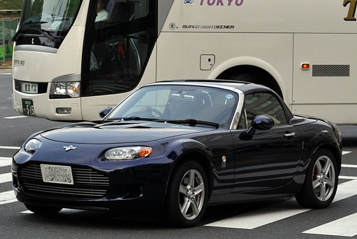 Model Aston Pistonheads - The image depicts a busy street scene in Japan. At the forefront, there's a black sports car with a distinctive hood ornament that resembles a shield, characteristic of its Volvo brand. The car is stationed near a white bus, which has a distinctive logo that reads "Super High Decker." The street is lined with white dashes, suggesting it might be a high-quality, well-maintained road. The presence of several people in the background suggests that the image was taken during the day in a populated urban area.