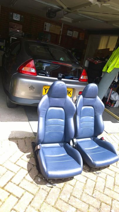 A group of people sitting on a bus in a parking lot - Pistonheads - The image depicts a garage with a large car parked inside. The car is predominantly silver. Nestled in front of the car, there are two blue automotive seats, placed as if they are part of the car they are replaced with, although they are not attached to anything and appear to be leaning sideways. In the background, there are items of various kinds, suggesting a common garage setup with tools and household items. The seats are positioned at an angle, with their backs facing the wall of the garage. The floor is adorned with a few leaves scattered around, indicating some activity outside the frame of the image. The overall scene implies a space used for both vehicle storage and possibly mechanical work or car interior installations, as suggested by the oriented seats.