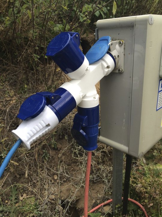 So who's getting an i3? - Page 59 - EV and Alternative Fuels - PistonHeads - The image shows a white garden hose with blue end attachments connected to a silver water meter, which is mounted on an electric box. The electric box stands on the ground, outside and surrounded by vegetation. The hose is anchored by a purple pole which appears to be set in the ground. The overall scene suggests a landscaping or gardening setup.