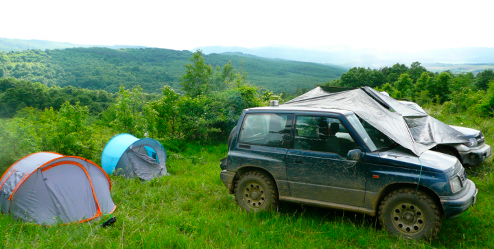 Little overnight trip to the Balkan range - Page 1 - Off Road - PistonHeads - The image shows a rugged outdoor scene, likely a camping site or a rest stop in a rural area. There is a small, suv-type vehicle parked in the center of the image, covered with a tarp, indicating it might be off-road or used for camping. Behind the vehicle, a tent is visible, which is a common element of such locations. Surrounding the tent and the vehicle, there is lush greenery, including trees and bushes, adding to the wilderness feel of the image. The background reveals hilly terrain under a clear, blue sky, further emphasizing the outdoor, nature-focused setting. The overall tone of the image suggests an adventurous and potentially remote location, commonly associated with camping and outdoor activities.