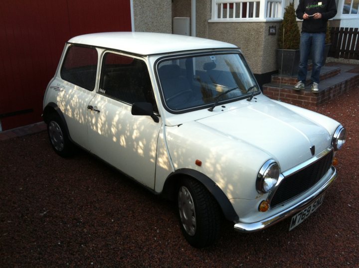 1995 mini sprite with 26000 miles - Page 1 - Classic Minis - PistonHeads - The image displays a vintage white car parked on a gravel surface next to a building. The car appears to be an older model, possibly from the 1960s or 1970s, with a classic design. The exterior of the car features a shiny metallic finish, indicating it might have been polished or cared for over the years. There are trees and greenery in the background, suggesting a suburban or semi-rural setting. A person is partially visible on the right side of the image, standing on a small staircase and observing the car. The overall lighting and shadows suggest that the photo was taken during daylight with ample sunlight, casting reflections on the car and giving the scene a bright and airy feel.