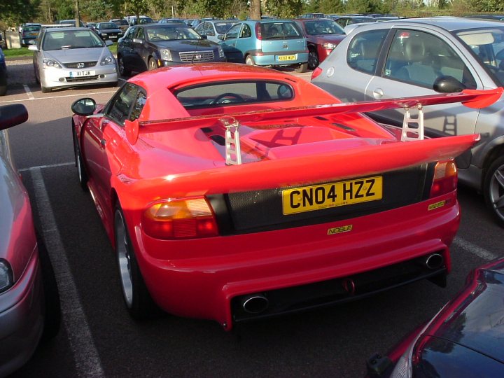 RE: Spoiler alert: PH Blog - Page 6 - General Gassing - PistonHeads - This is a photograph taken at an outdoor parking area. In the foreground, the main focus is on a vibrant red sports car with its rear hatch open, displaying a large rear wing. The license plate reads "CND4 HZZ" with a small decal that says "NAUGLE" below it. This car is parked among several other vehicles of various colors, primarily metallic. The lighting suggests an overcast day, and the angle of the shot gives a side view of the car, highlighting its sleek lines and the unique design of the rear spoiler wing.